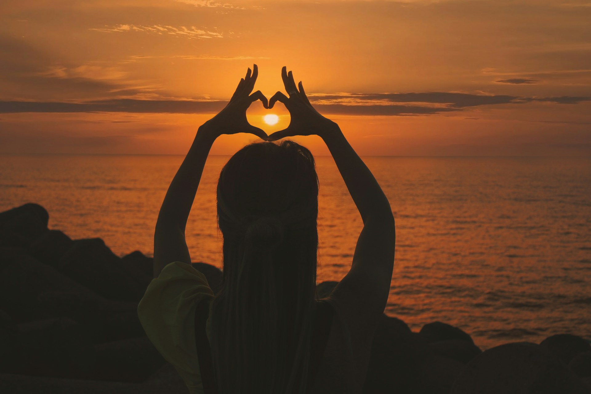 Person making the shape of a love heart in front of the setting sun